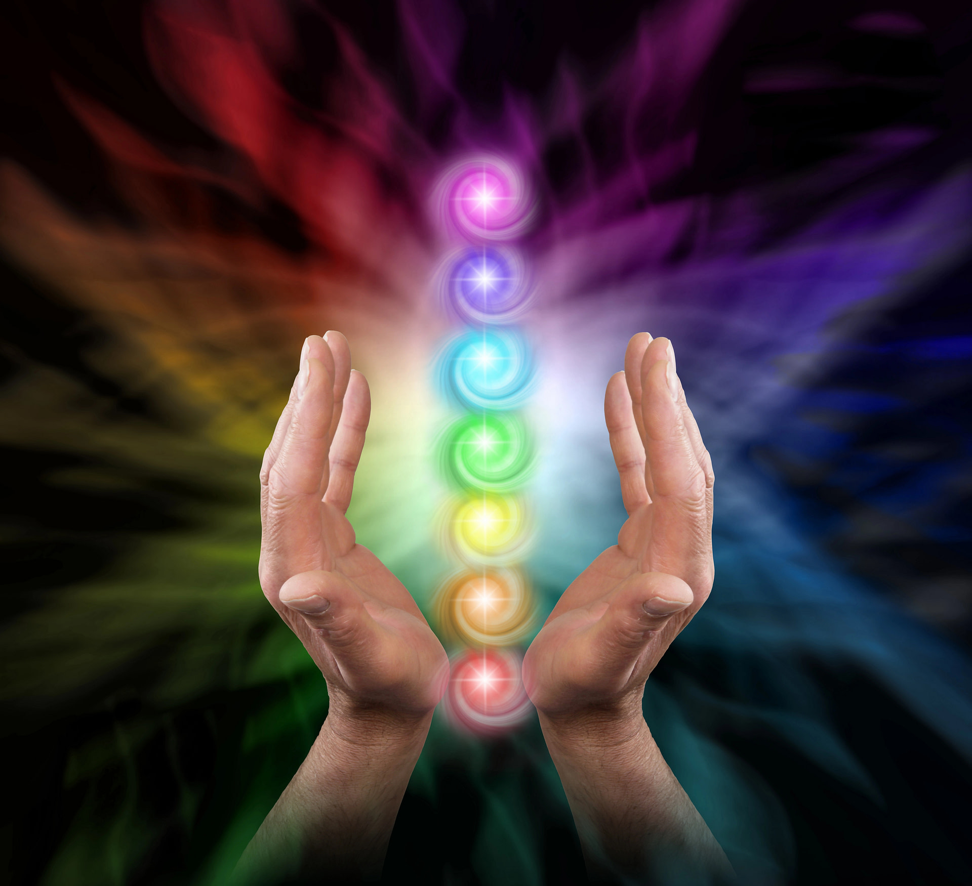 Sending chakra healing energy - male parallel hands facing upwards against a multicoloured background of energy and the seven chakras floating between hands